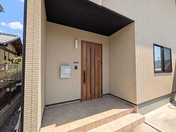 木目調の玄関ドアが外観に調和しています。広々としたタイルのアプローチが特徴です。屋根付きのポーチで雨の日も安心して出入りできます。シンプルなデザインが周囲と調和しています。