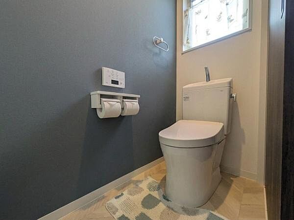 落ち着いた色合いの壁が印象的なトイレです。小窓から自然光が入り明るさを感じられます。ダブルペーパーホルダーが設置されていて便利です。シンプルな内装で清潔感があります。