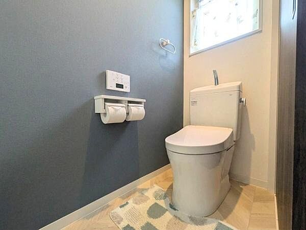 落ち着いた色合いの壁が印象的なトイレです。小窓から自然光が入り明るさを感じられます。ダブルペーパーホルダーが設置されていて便利です。シンプルな内装で清潔感があります。