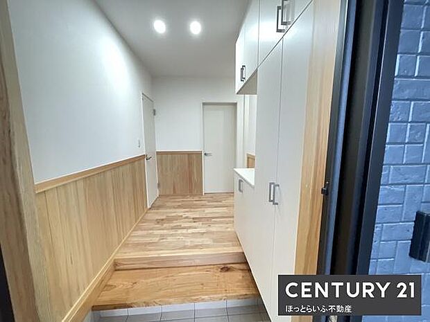 明るい照明が設置されている玄関です。木目調の腰壁と床が温かみのある印象を与えます。収納スペースも充実しておりすっきりとした空間です。段差が明確で使い勝手が良いデザインです。