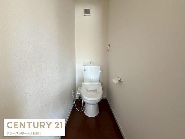 シンプルなデザインのトイレです。壁と床の色合いが落ち着いた印象を与えます。換気設備が設置されています。ペーパーホルダーやコンセントなどの設備も整っています。