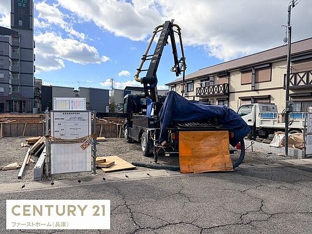 建築中の現場がしっかりと管理されています。資材や重機が整然と配置されており作業の様子がわかります。街並みに調和した周辺環境も確認できます。天候にも恵まれており明るい雰囲気です。