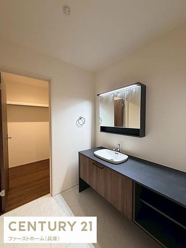 木目調の洗面台とブラックのカウンターが空間に落ち着きをもたらします。ミラーキャビネットで収納も充実しています。明るい照明が印象的な洗面所です。奥には便利なスペースが設けられています。