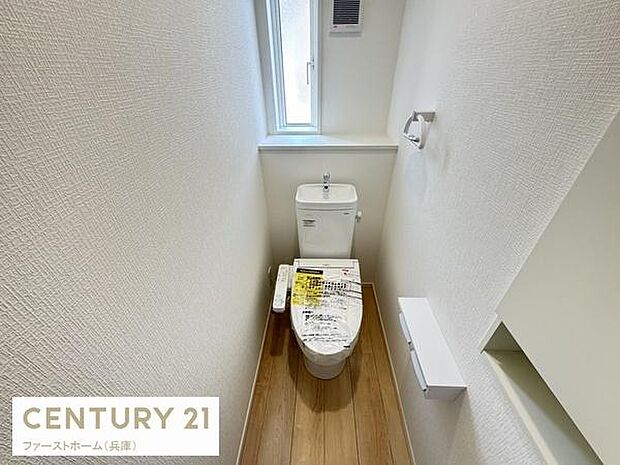 明るい窓付きで自然光が入る快適なトイレ空間です。清潔感のある白い壁と落ち着きのある木目調の床が調和しています。ペーパーホルダーや収納も備わっており使い勝手にも配慮されています。