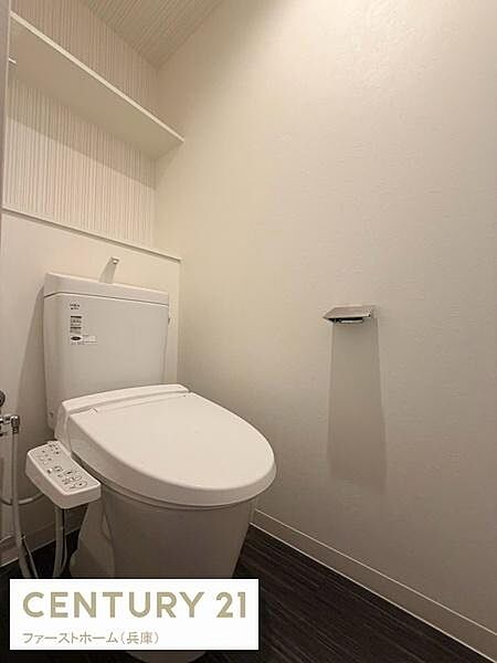 清潔感のある内装が印象的なトイレです。壁面の棚が備わっており収納スペースも確保されています。温水洗浄機能付きで使い勝手にも配慮されています。床材や壁紙の色合いも落ち着いています。