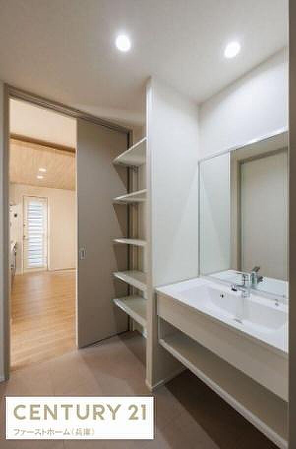 明るい照明が印象的な洗面台で清潔感のある空間です。使いやすい大きめのミラーが設置されています。オープンタイプの収納棚が備え付けられており整理整頓もしやすい設計です。廊下からのアクセスも便利な位置です。