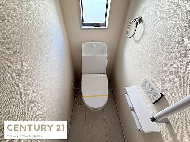 コンパクトな空間に設置されたトイレです。窓付きで換気もしやすい設計となっています。清潔感のある壁紙と床材が使用されています。ペーパーホルダーや手すりも完備されています。温水洗浄便座タイプのトイレです。