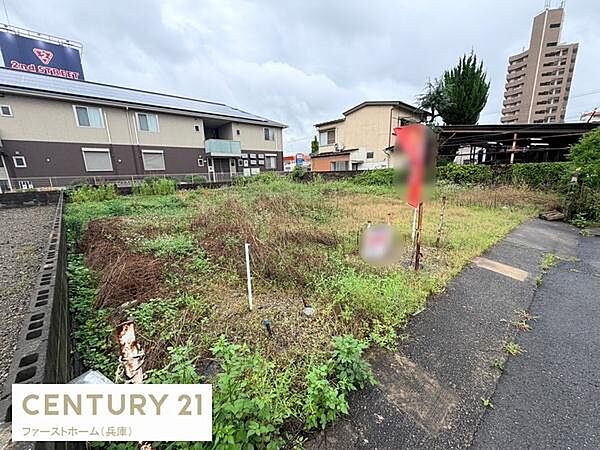 現地土地写真です。周囲には住宅や建物が建ち並びアクセスしやすい舗装された通路が設けられています。敷地は開放的で様々な活用が可能です。草木が生い茂っており現状を確認いただけます。