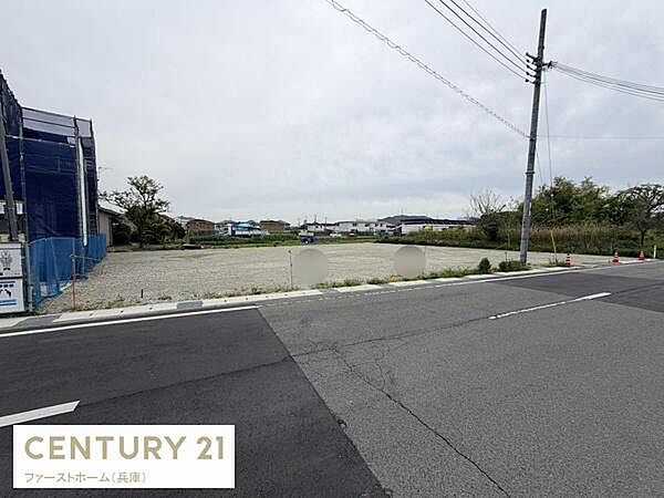 舗装された道路に面した現地土地写真です。周辺には建物や緑も見え広々とした空間が確保されています。電柱やインフラも整っており様々な用途に対応しやすい環境が整っています。
