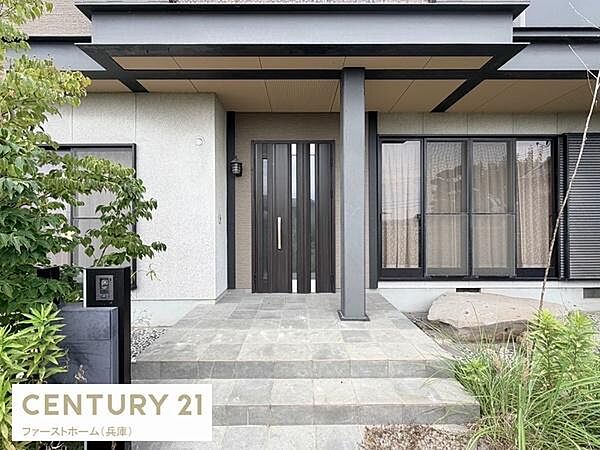 玄関はシンプルなデザインで広々としたタイル敷きのアプローチが印象的です。大きな窓が隣接し明るい印象を与えます。外観の植栽がアクセントになっています。重厚感のある扉が特徴的です。
