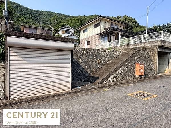 前面道路に面した現地土地写真です。高台に位置し周辺は落ち着いた住宅エリアです。敷地内には既存のガレージや階段が設けられています。傾斜を活かした土地形状が特徴的です。周囲の緑も心地よい環境です。