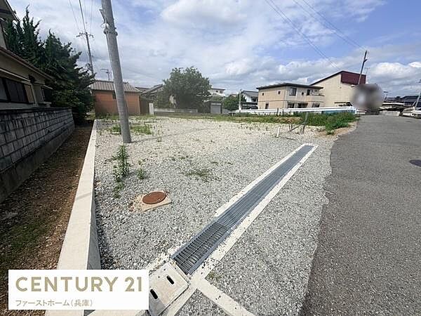 周辺には落ち着いた雰囲気の住宅が並び道路に面した現地土地写真です。敷地には砂利が敷かれており整った形状が印象的です。前面道路は広めで車両の出入りもスムーズに行えます。電柱やインフラ設備も整っています。
