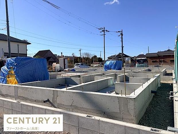 現地からは周囲の住宅街や空のひらけた眺めが広がっています。晴れた日の青空が印象的です。建物の基礎工事の様子が確認できます。周囲には静かな住宅が並んでいます。