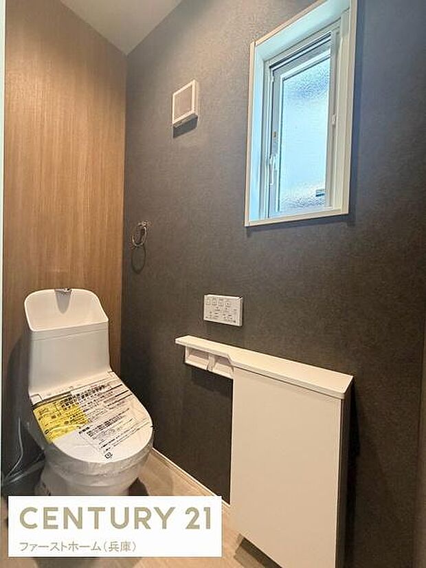 落ち着いた色合いの壁紙と木目調のアクセントが特徴のトイレです。手洗い付きのトイレで使いやすさも考えられています。窓があるため換気しやすく快適な空間が保てます。収納スペースもあり機能性が充実しています。