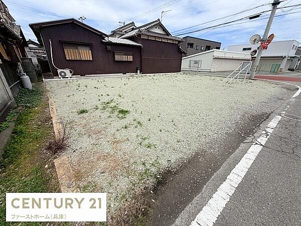 道路に面した現地土地写真です。周囲は住宅や施設が点在し落ち着いた環境がうかがえます。敷地は整った形状で利用しやすい印象があります。日当たりの良さも感じられる現地土地写真です。