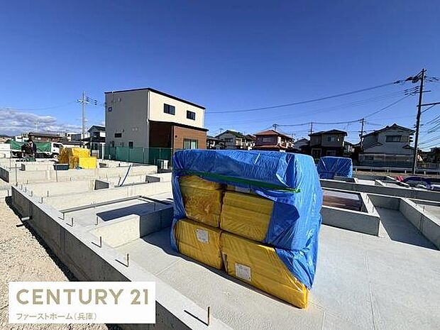 青空の下でしっかりとした土台工事が進行中です。周辺には住宅が立ち並び安心感のある街並みです。建築資材も整然と保管され現場の管理が行き届いています。基礎部分が確認できる状態で建築の進捗が明瞭です。