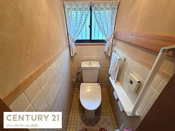 窓付きで自然光が入る明るいトイレです。壁には手すりや収納棚があり利便性を高めています。落ち着いた色合いのタイルが空間に統一感を与えています。カーテン付きの窓でプライバシーも確保できます。