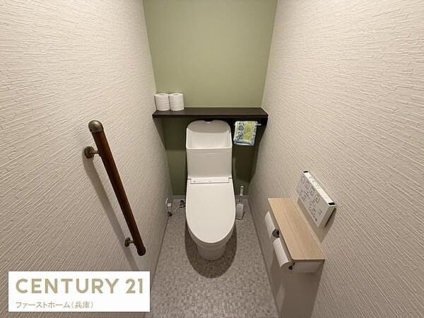 ナチュラルな色合いの壁が印象的なトイレです。手すりが設置されており安心してご利用いただけます。収納棚には小物やペーパーを置くことができて便利です。温水洗浄機能付きで快適にお使いいただけます。
