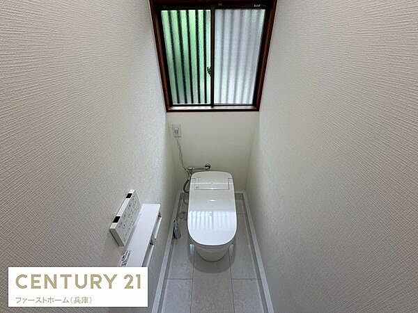 窓があるため自然光が入りやすく明るい印象のトイレです。壁紙や床材はシンプルで清潔感があります。温水洗浄便座が設置されています。ペーパーホルダーやリモコンも壁付けで配置されています。