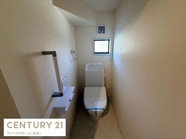 コンパクトな空間に設置されたトイレです。小窓があり換気や採光に配慮されています。ペーパーホルダーやタオル掛けも備わっています。壁紙や床材は落ち着いた色合いです。清潔感のあるシンプルなデザインです。