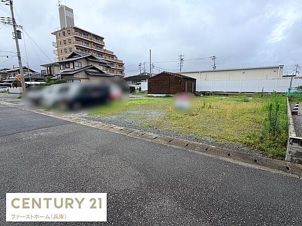 道路に面した現地土地写真です。周辺には住宅や建物が立ち並んでおり落ち着いた環境が特徴です。整形地で利用しやすい形状になっています。前面道路も広くアクセスが良好です。