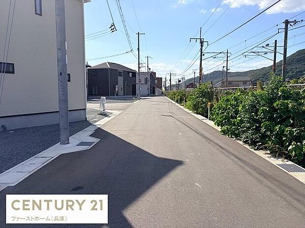 前面道路は幅が広く車の出し入れがしやすい印象です。周囲には電柱や緑が配置されており開放感があります。建物の外観も現代的で清潔感が感じられます。空の広さが印象的な現地写真です。