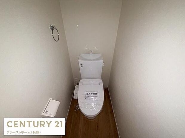 シンプルで清潔感のあるトイレ空間です。壁紙や床材も落ち着いた色合いで統一されています。手洗い器やペーパーホルダーも設置されています。全体的にゆとりのあるスペースです。