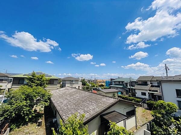 抜けるような青空と白い雲のコントラストが美しく、開放的な眺望です。陽光を浴びた緑豊かな木々と立ち並ぶ家々が調和し、穏やかで平和な日常の空気感を存分に味わうことができます。※AIによる画像加工あり
