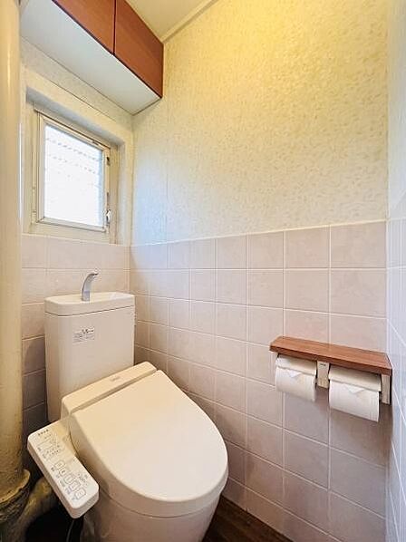 窓からの自然光で明るく、清潔感のあるトイレです。お手入れしやすいタイル張りの壁や、小物を置ける便利な棚付き2連ペーパーホルダーが魅力。上部の吊り戸棚で収納スペースもしっかり確保された快適な空間です。