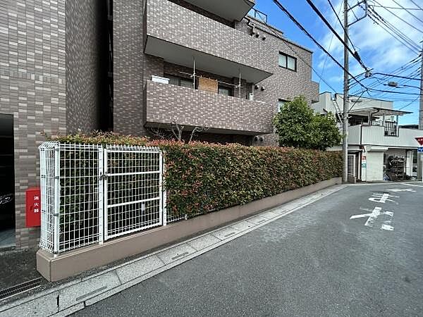 前面道路から見た、マンションの外観です。茶系のタイル張りの外壁で、道路沿いには生垣とフェンスが続いています。オートロック、エレベーター、宅配ボックスを備えています。