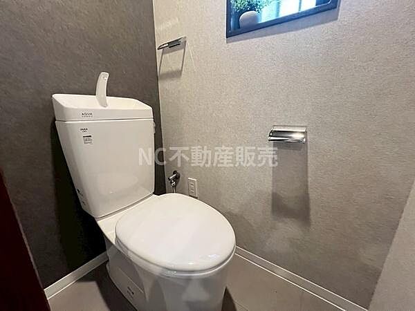 シンプルで使いやすいトイレ空間です。白とグレーの落ち着いた色合いが特徴です。ペーパーホルダーやタオル掛けが設置されています。窓から光が差し込みます。清掃もしやすい設計です。