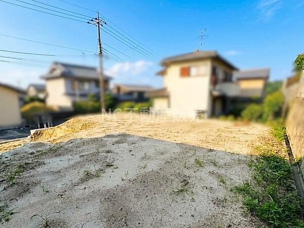 周辺に住宅が立ち並ぶ現地土地写真です。陽当たりが感じられる開放的な雰囲気が広がっています。整地されており前面道路との接道も確認できます。電柱や電線の位置もわかりやすく周囲の環境も視認できます。