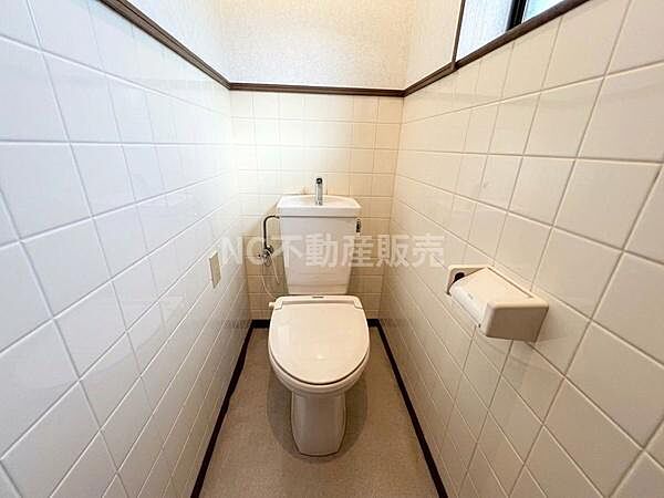 白を基調としたタイル張りの壁が清潔感を感じさせるトイレです。上部に窓があり明るさも確保されています。手洗い付きタンクを採用しており使い勝手も良好です。