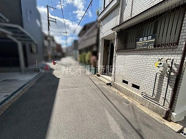 前面道路は舗装されており周囲には住宅が立ち並ぶ環境です。物件は白いタイルの外観が特徴的です。道路幅もあり車の通行も可能です。電柱や配管なども整備されています。落ち着いた雰囲気の街並みが見られます。