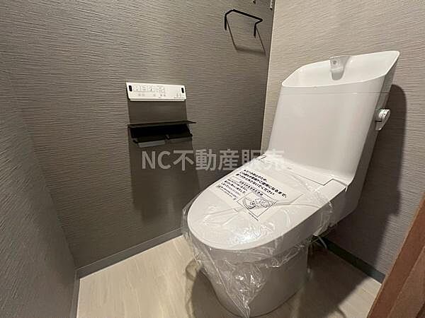 シンプルで清潔感のあるトイレ空間です。落ち着いた色合いの壁紙が印象的です。ペーパーホルダーや棚もすっきりと配置されています。手洗い一体型で使いやすさが考慮されています。床材も掃除がしやすい仕様です。