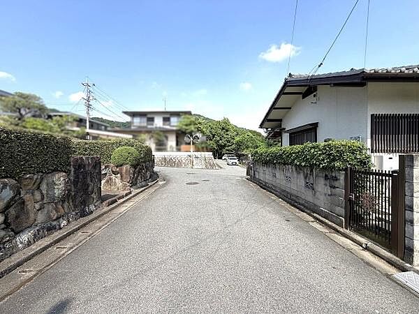 北側幅員約6mの前面道路。ゆとりある道幅と整備された街並み。両側に石積みや生垣が続く、閑静な住宅地の雰囲気が伝わります。遠景には山も望めます。
