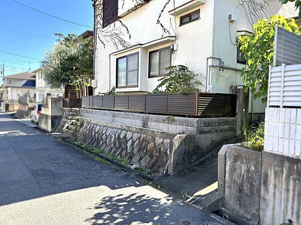 現地土地写真では道路に面した立地が確認できます。石垣やフェンスが設けられており敷地の境界がわかりやすいです。建物周辺には緑も見られ落ち着いた街並みが感じられます。アクセスしやすい環境です。