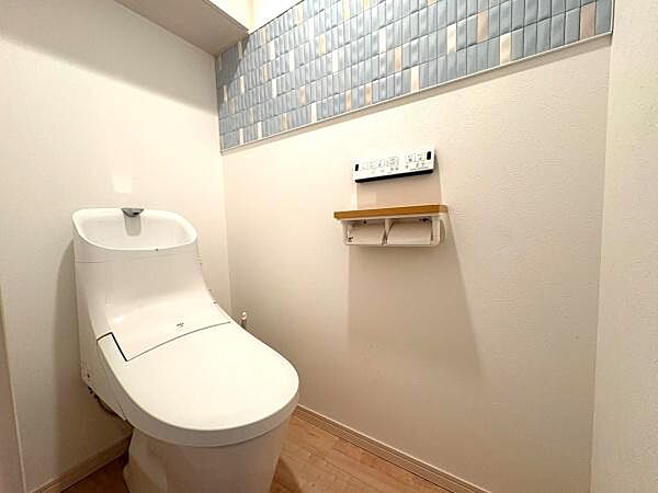 清潔感のあるホワイトのトイレ本体と淡いブルータイルのアクセント壁が落ち着いた空間を演出しています。壁面のリモコンや棚も配置され使い勝手にも配慮されています。