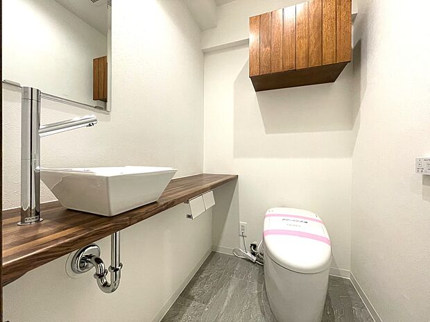 タンクレスですっきりとしたデザインのウォシュレット付きトイレです。室内は天然無垢材の温もりを感じる落ち着いた上質な空間です。吊戸棚も設置されています。ミラー付の便利な専用手洗いスペースも付いています。